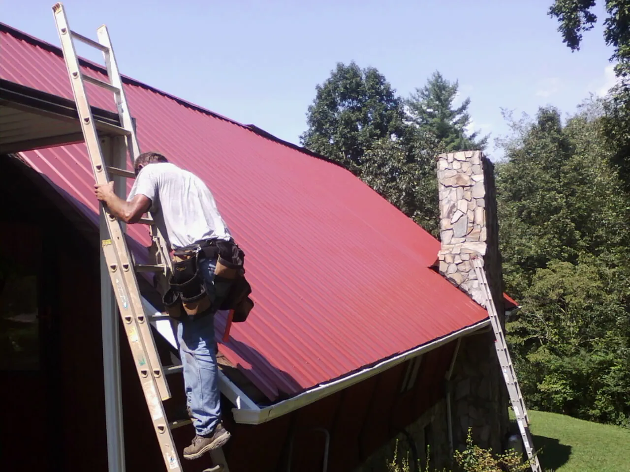 Roofer on a ladder beside a red metal roof and stone chimney — Soffit & Fascia Repair by Roofing Brothers in Roseburg
